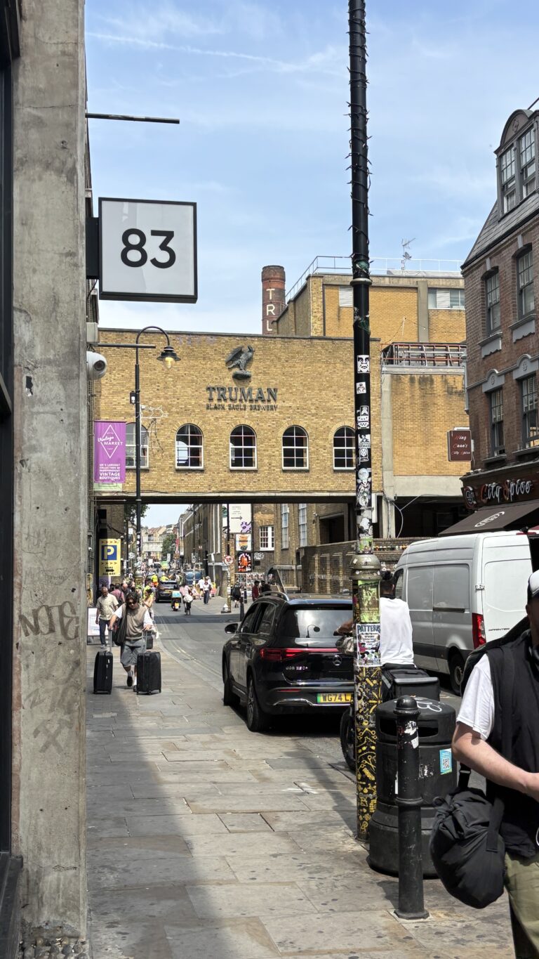 Historic Truman Brewery building in Shoreditch, East London, with its iconic chimney and red brick facade, set against a clear sky, surrounded by street art and urban surroundings.