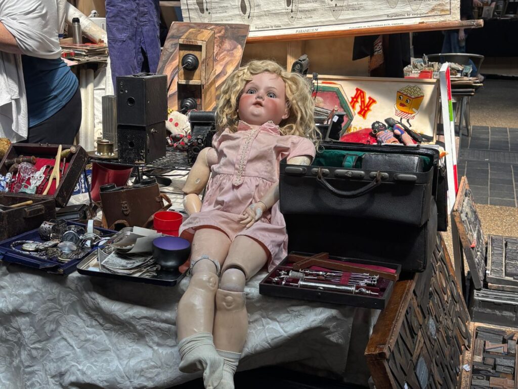 Antique child's doll in pink dress at an antique dealers store in Old Spitalfields Market, East London surrounded by other antiques and trinkets.
