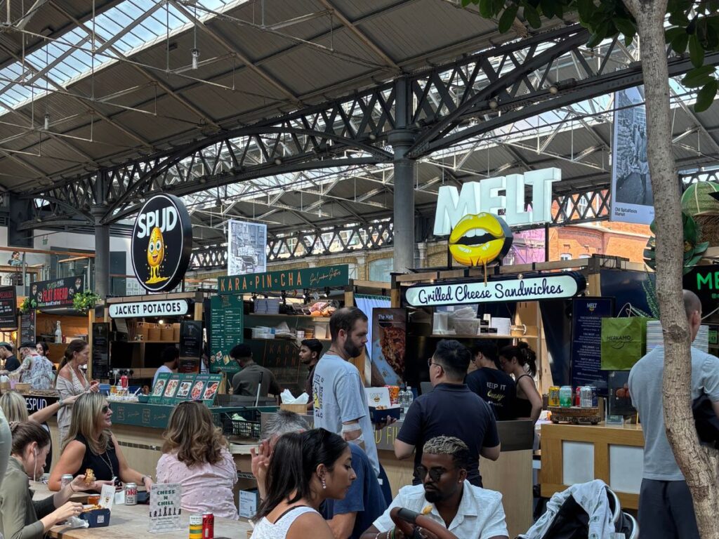 Food stalls at Shoreditch's Spitalfields Market with diners sitting in front in a casual setting.