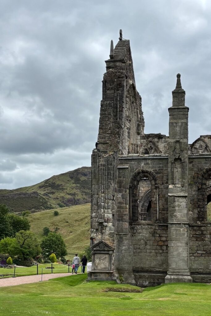 Ruins of Holyrood Abbey at the Palace of Holyroodhouse surrounded by green landscape under cloudy skies.