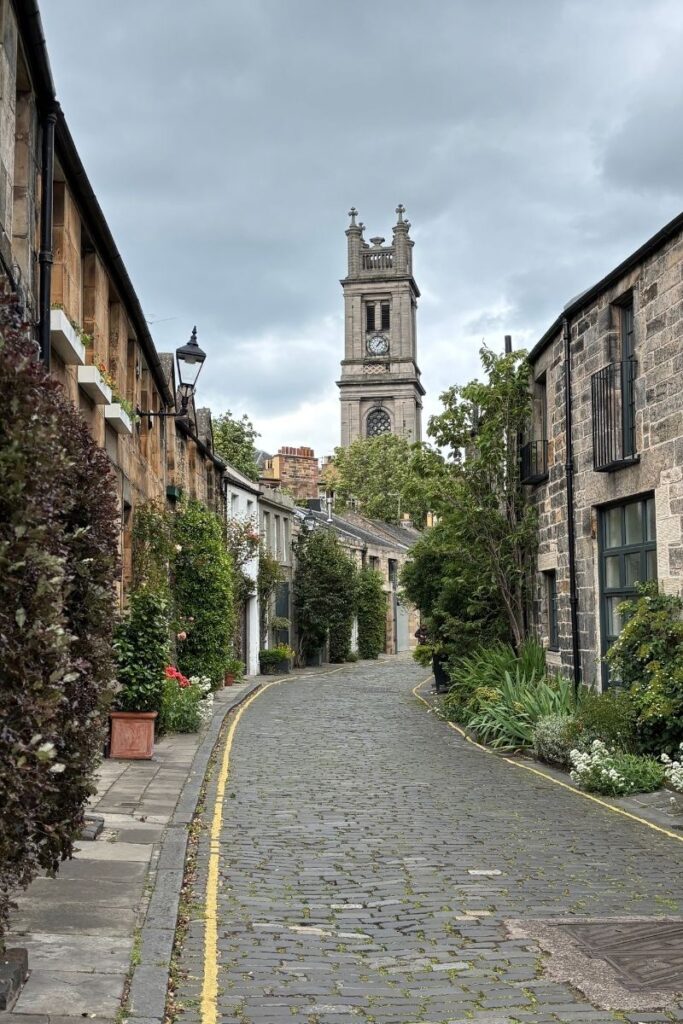 Cobbled Circus Lane in Stockbridge, Edinburgh, lined with stone mews houses and flower boxes, with St Stephen’s Church tower visible in the background.