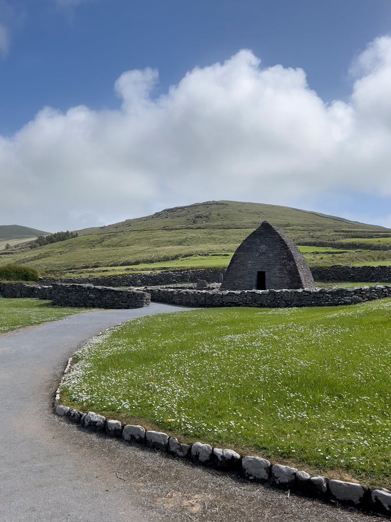 Stone building in Dingle, Ireland with green landscape and hill under a cloudy sky.