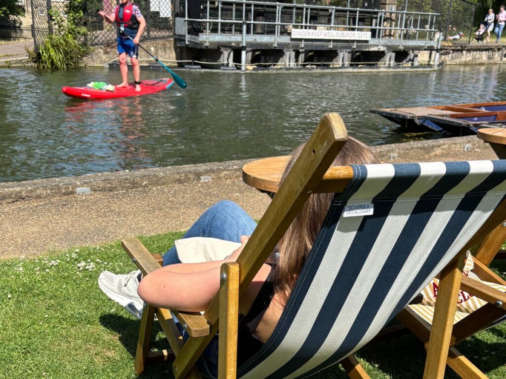 Person relaxing in a black-and-white striped deck chair facing a calm river, while another person paddles by on a paddle-board.