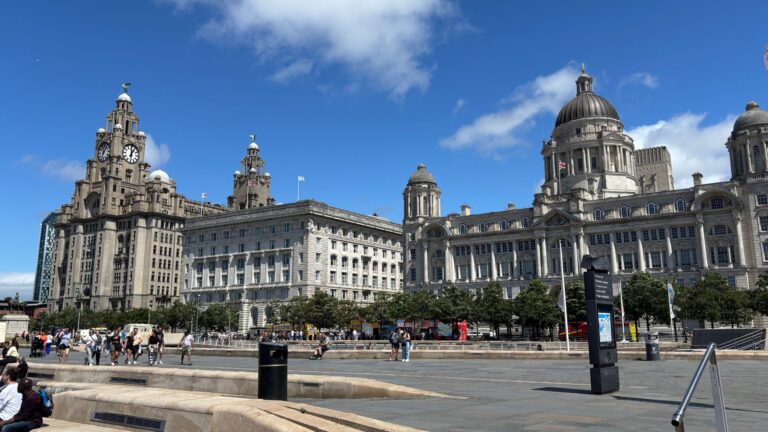 Liverpool waterfront with the Three Graces—Royal Liver Building, Cunard Building, and Port of Liverpool Building—framed against a clear sky as seen from across the Pier Head.