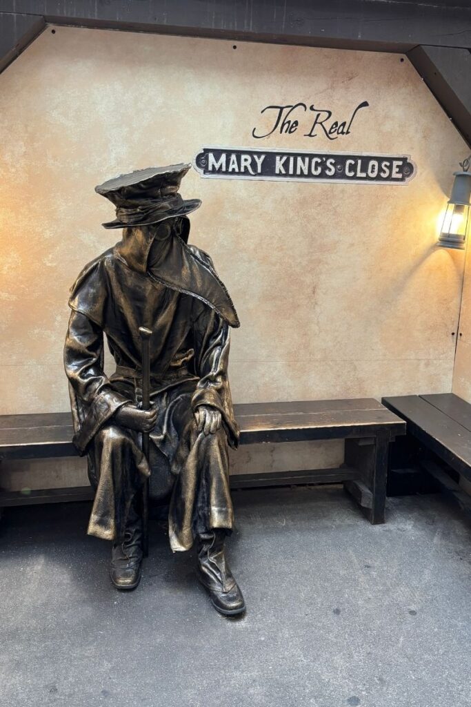 Statue of a plague doctor with beak-shaped mask and cloak sitting on a bench outside The Real Mary King’s Close in Old Town Edinburgh.