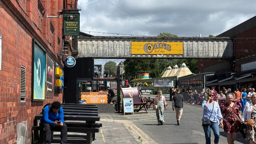 Baltic Triangle Village in Liverpool with bridge across connecting old brewery buildings and yellow Cains signage. Two people walking along pedestrianized street.