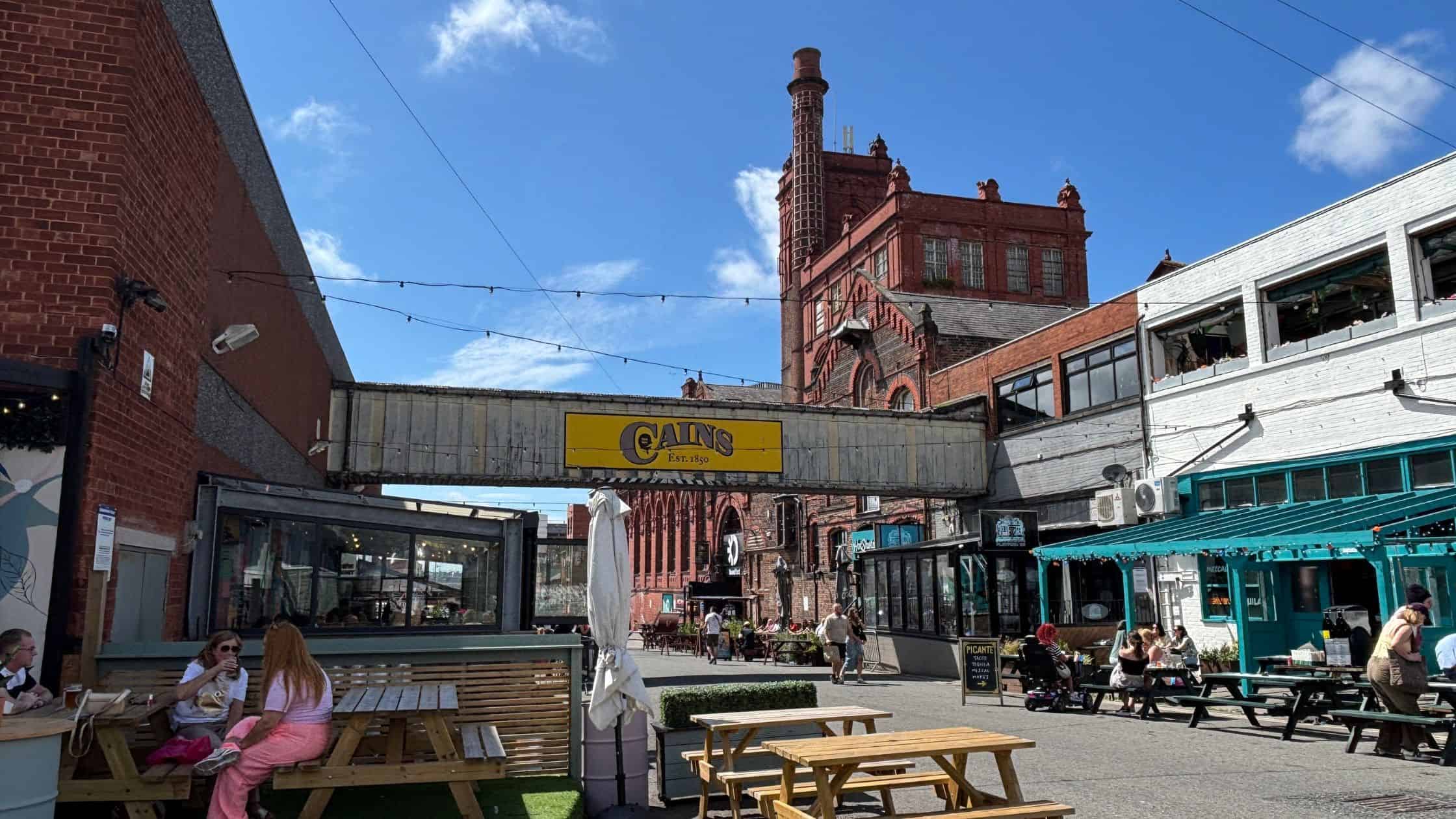 Baltic Triangle Liverpool Cains Brewery Village with chimney of original image in background and bars with outdoor seating.