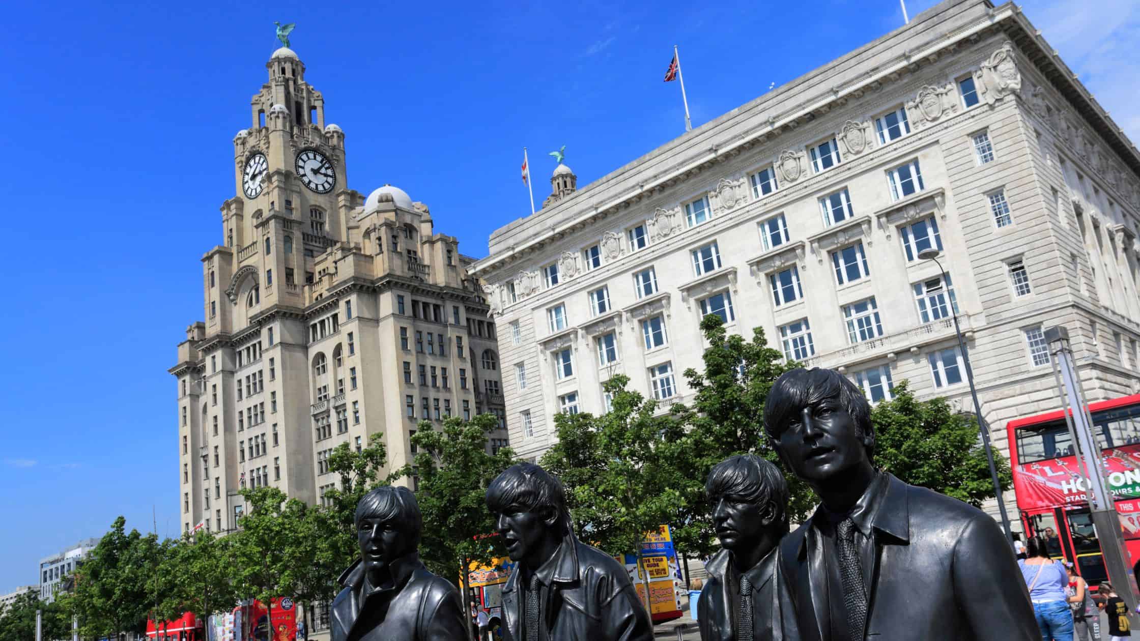 Famous Liver Building on Liverpool's waterfront against a blue sky with statue of The Beatles in front.