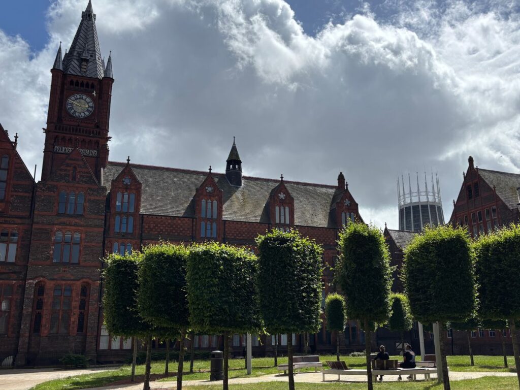 Red brick Victorian Museum & Gallery building in Liverpool's Knowledge Quarter with iconic clock tower on left and circular Liverpool University green space with trees in foreground.