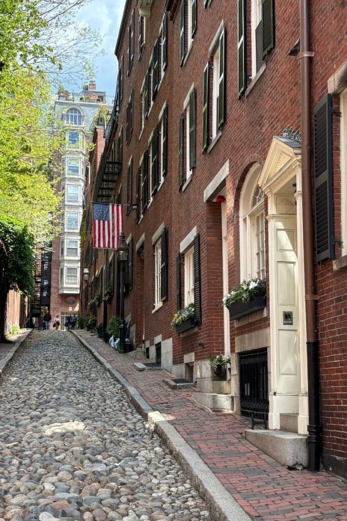 Streetscape looking up cobbled Acorn Street in Beacon Hill Boston with federal style homes on the right and a hanging American flag.