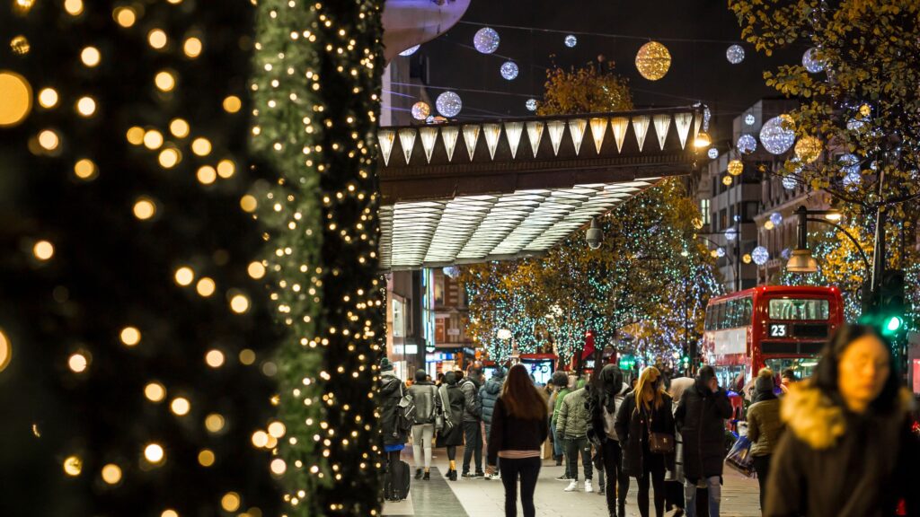Christmas lights illuminating Oxford Street in London at night, with festive shop windows and busy holiday shoppers.