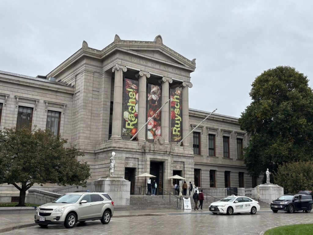 Front entrance of the Museum of Fine Arts, Boston on a cloudy, rainy day with wet pavement and cars parked outside.
