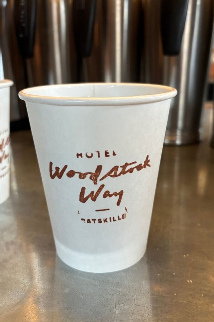 White paper coffee cup with words "Hotel Woodstock Way" printed in brown lettering on the front sitting on a wooden countertop.