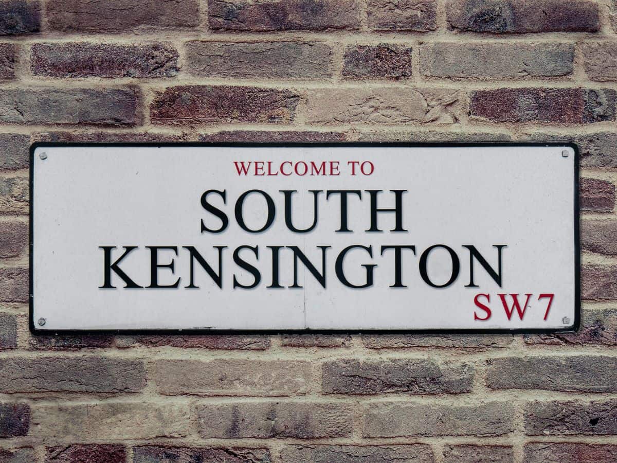 White street sign with words South Kensington in capital letters against brick wall.