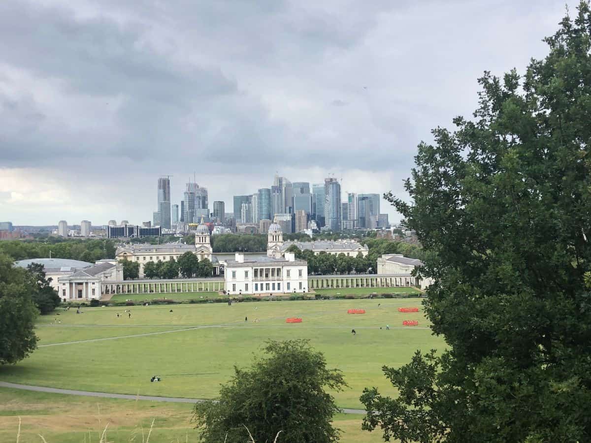 London skyline as seen from Royal Observatory including Maritime Museum in foreground and Canary Wharf in the distance against a cloudy sky.