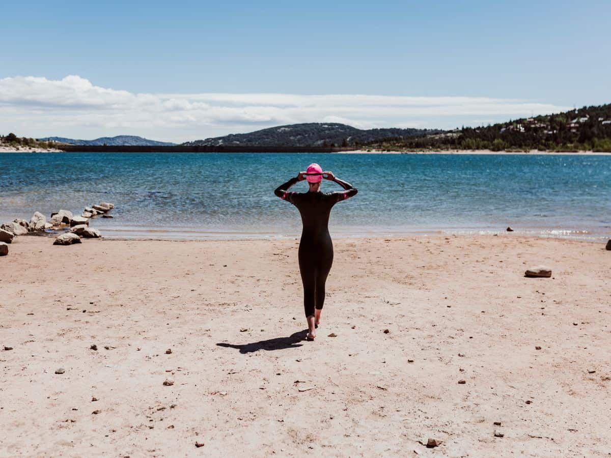 Women in wetsuit and pink swimming cap standing on a beach looking out to blue waters anticipating a swim