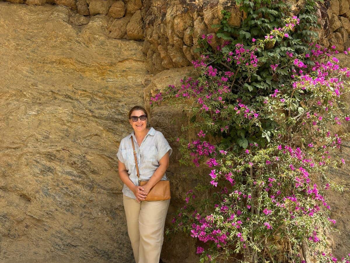 Middle aged women dressed in light summer clothes standing against a wall built into the rock in Barcelona's Parc Guell with flowers on the right.