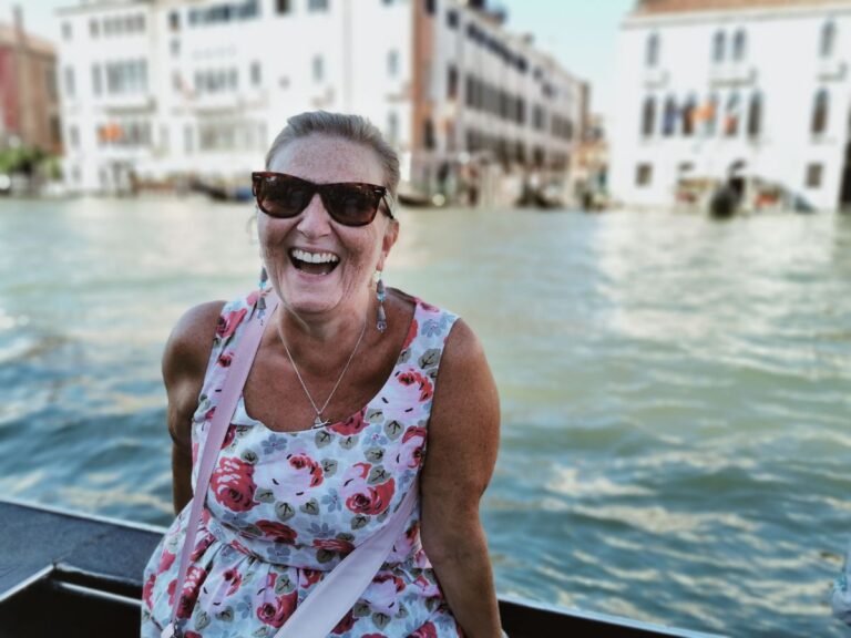 A senior woman smiling happily in Venice with water and historic architecture in the background.