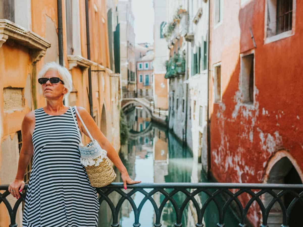 Elegant senior woman in striped dress posing by a canal in Venice, capturing the essence of European style