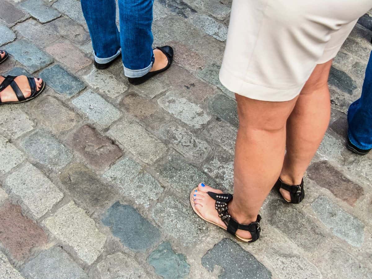 Middle-aged people's legs standing on cobblestones. Three people are wearing sandals and one is wearing shorts.