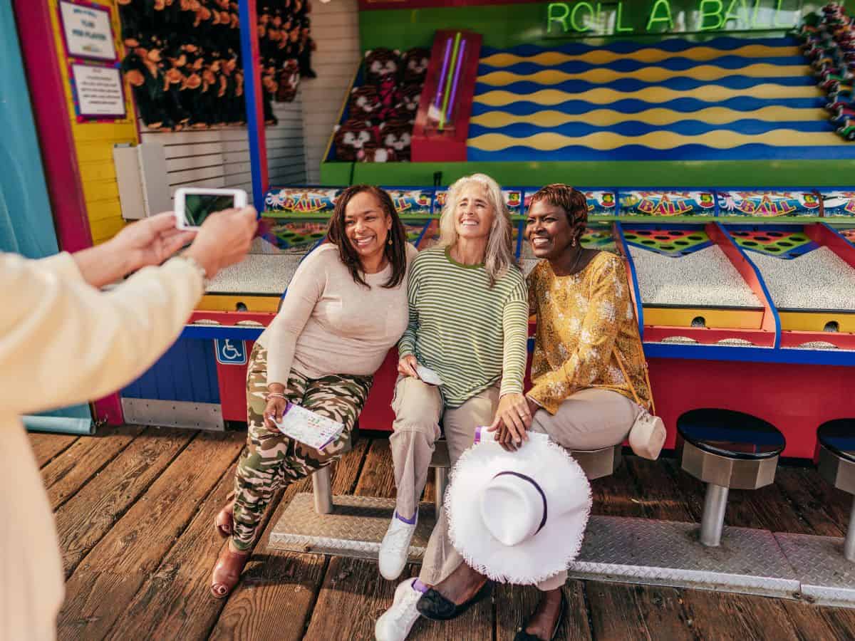 Three friends over 50 sitting on a bench having their photo taken. One women his holding a white hat.