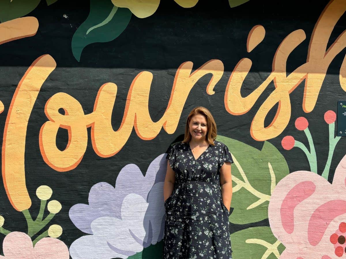 Midlife woman in floral dress standing in front of colorful street mural of flowers and the word "Flourish"